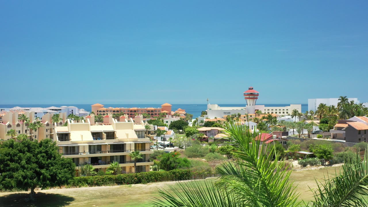 Elevated view over Los Cabos with cream buildings, palms, open ground, a red-topped tower, and the ocean horizon under clear blue sky, no visible people.