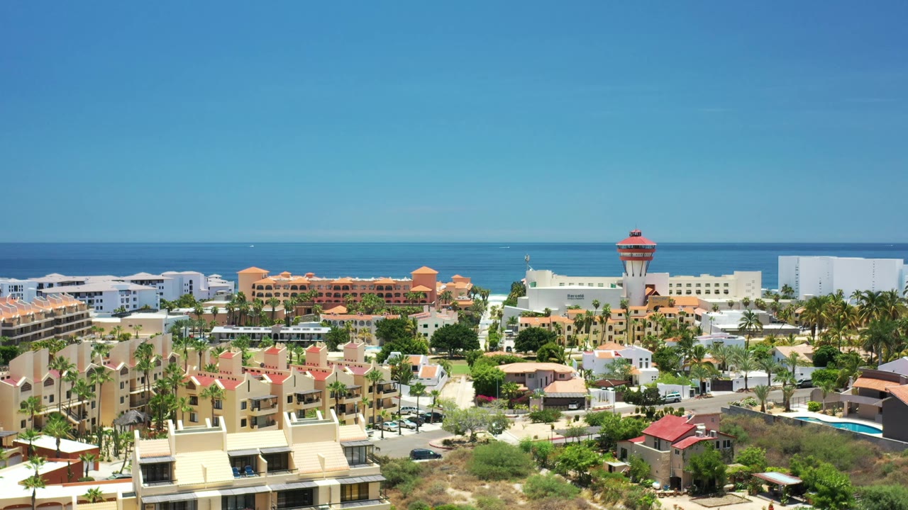 Aerial frame in Los Cabos showing cream villas, palm rows, white hotel blocks, and the ocean stretching behind them, with no visible people.