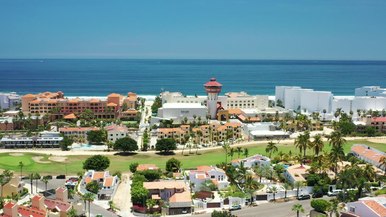A wide aerial view over Los Cabos, Mexico shows blue ocean, white surf, resort buildings, orange rooftops, palms, roads, and no visible people at the start of the clip.