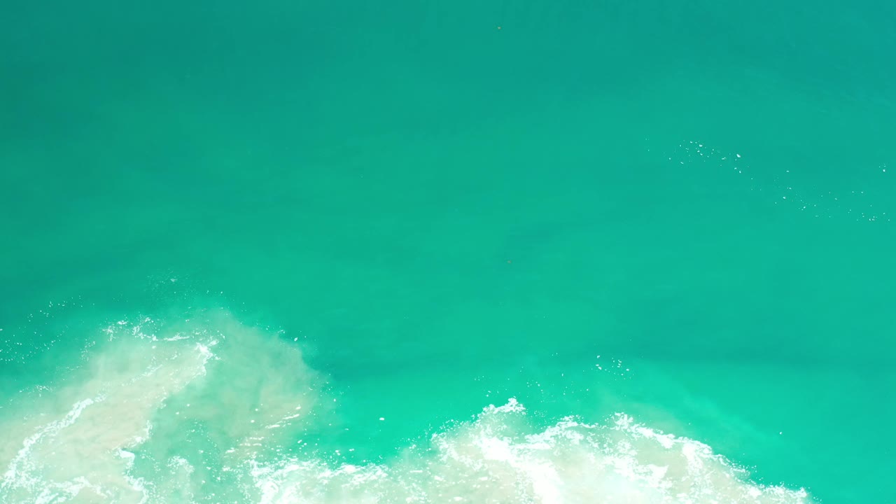 Overhead view of clear turquoise ocean water in Los Cabos, Mexico, with white shore foam entering along the lower edge and no visible people.