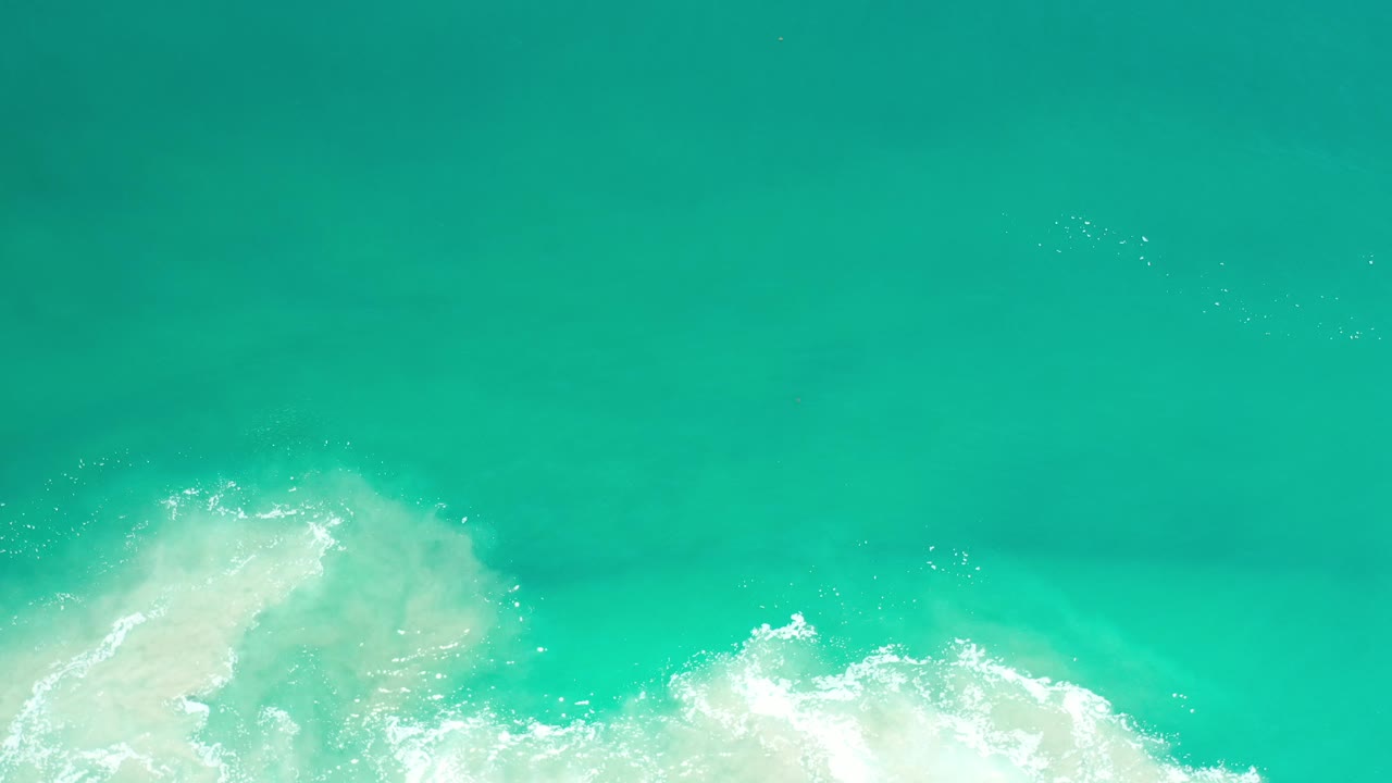 Overhead Los Cabos ocean view with smooth turquoise water above and white surf foam pushed farther across the bottom of the frame, with no visible people.