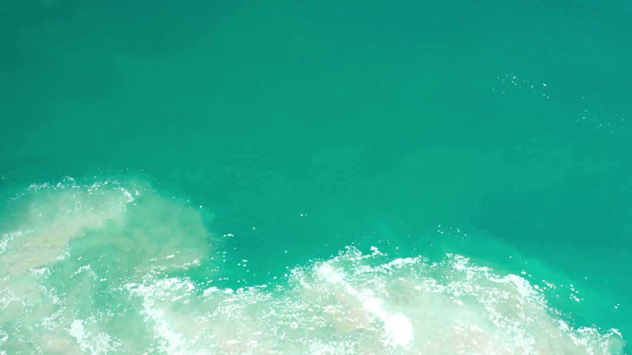 Overhead view of bright green ocean water in Los Cabos, Mexico, with white foam entering from the bottom and no visible people.