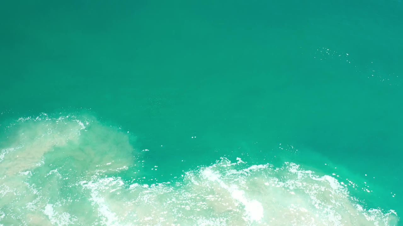 Aerial-looking frame of Los Cabos green ocean water with churning white foam, pale murky sand, surface sparkle, and no visible people.