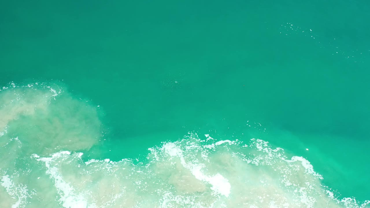 Aerial Los Cabos water view with a broad pale reef-like patch under the surface and white foam spreading across the lower half, no visible people.