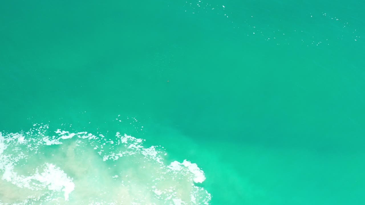 Top-down view of turquoise sea in Los Cabos, Mexico, with a band of white surf entering from the lower left and no visible people.