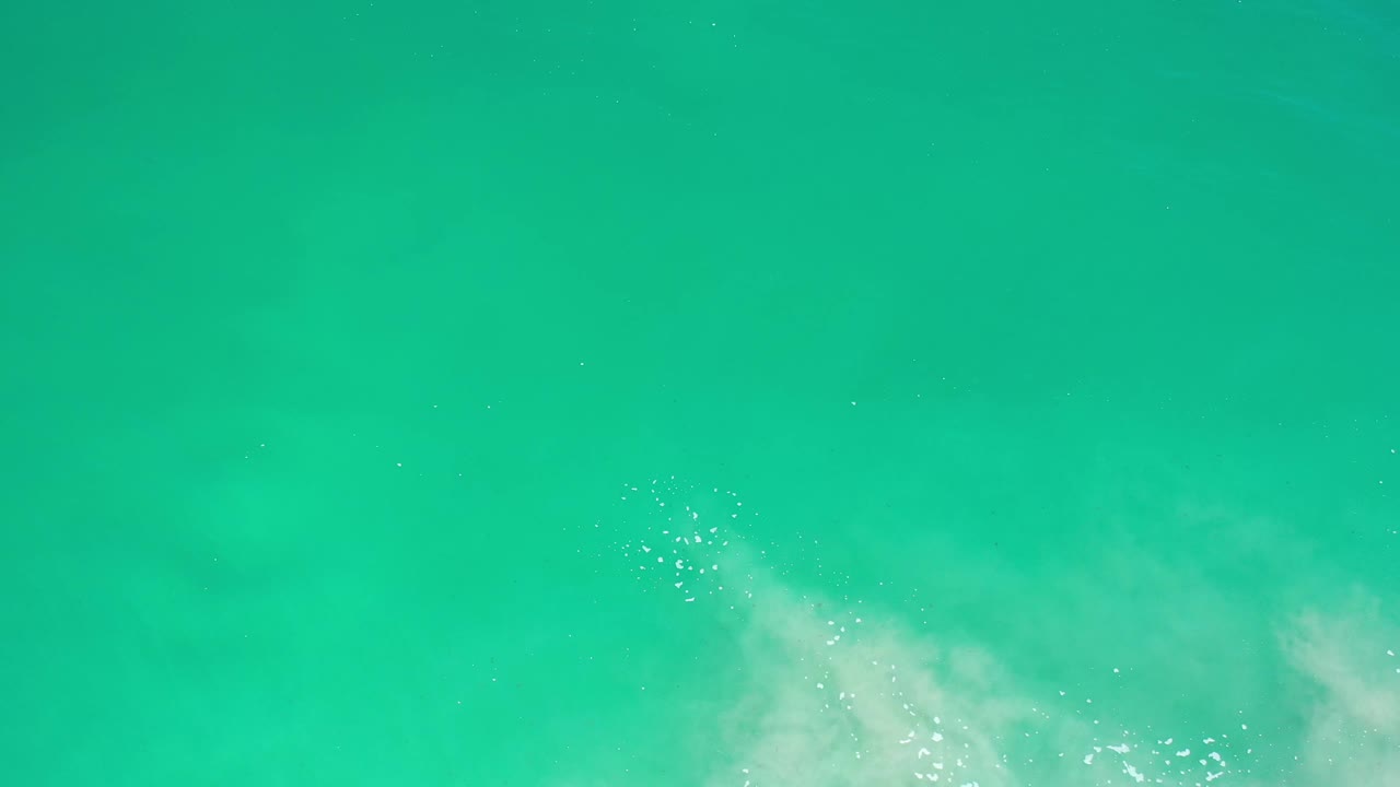 Overhead Los Cabos water frame with turquoise-green surface, clustered sunlight glints, and a soft white foam patch rising from the lower center, no visible people.
