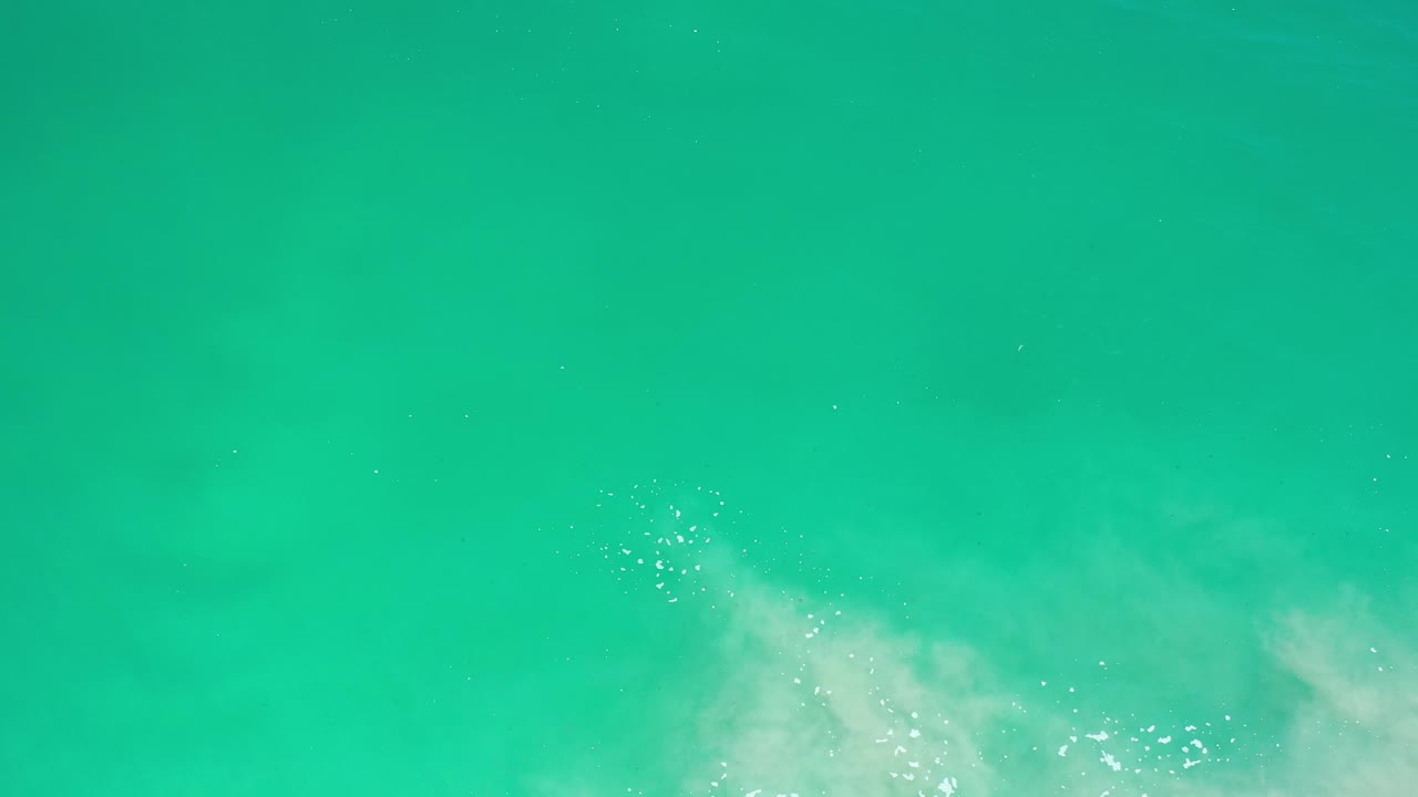 Top-down view of green coastal water in Los Cabos, Mexico, with small bright reflections and white wave foam concentrated along the lower right edge, no visible people.
