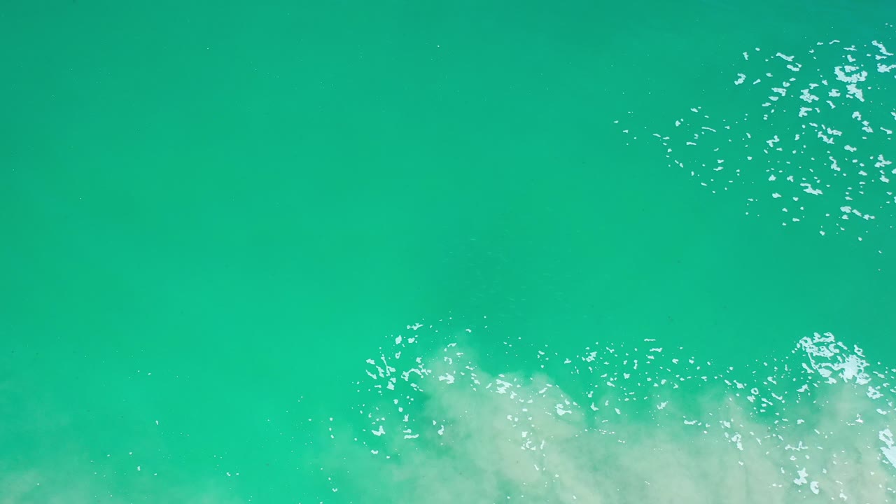 Overhead view of green ocean water in Los Cabos, Mexico, with a soft white foam mass entering along the lower edge and no visible people.