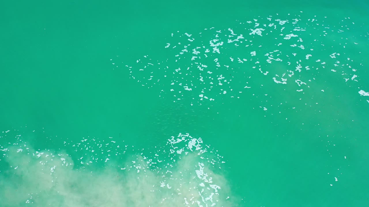 Overhead view of turquoise ocean water in Los Cabos, Mexico, with white sun flecks and pale foam entering from the lower edge, no visible people.