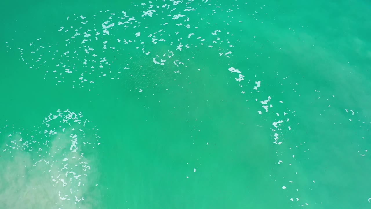 Overhead Los Cabos ocean frame with bright white flecks across green-blue water and a pale cloudy patch near the lower edge, no visible people.