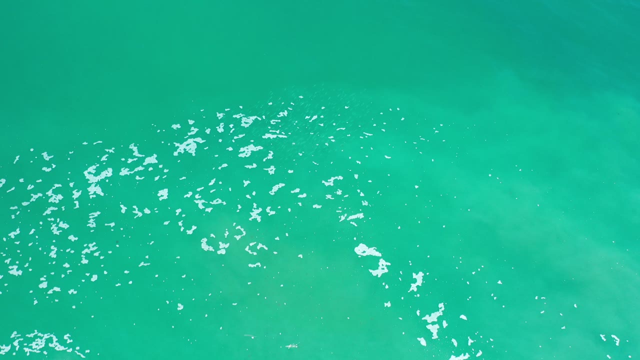 Blue-green ocean water in Los Cabos, Mexico, seen from above with white foam clustered across the lower left and no visible people.
