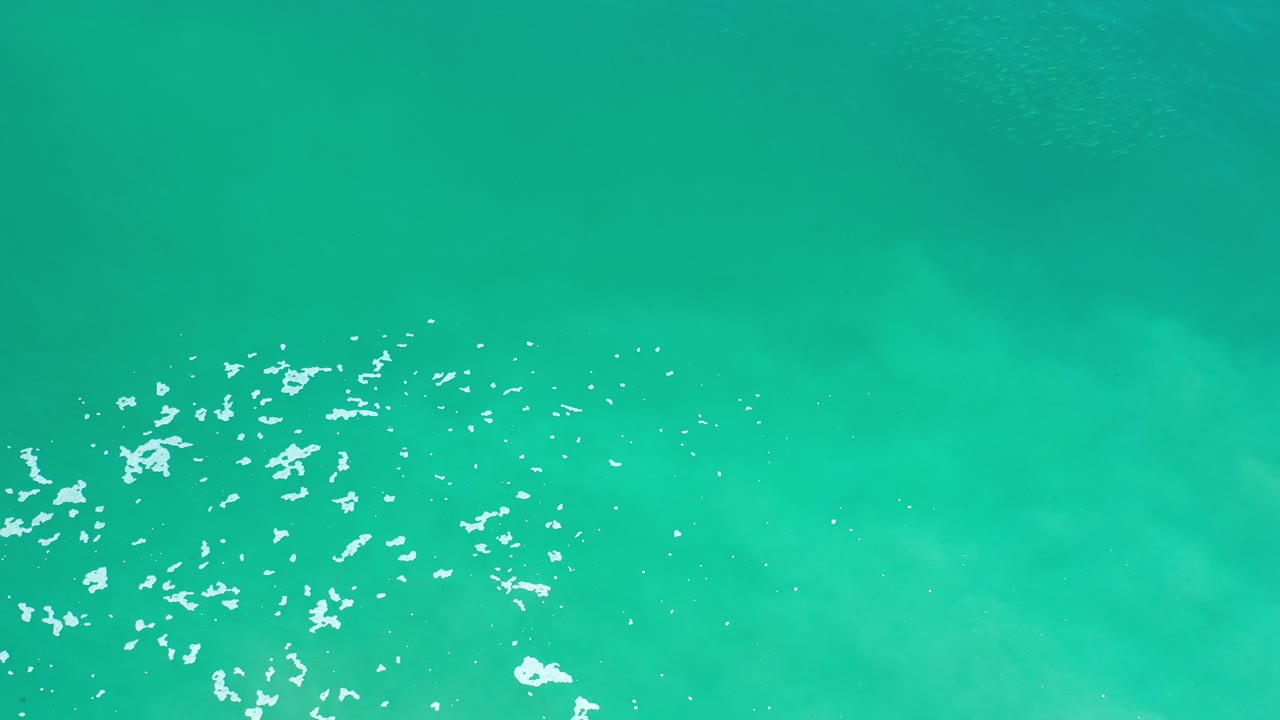 Top-down view of blue-green ocean water in Los Cabos, Mexico, with a cluster of white foam entering the lower left and no visible people.