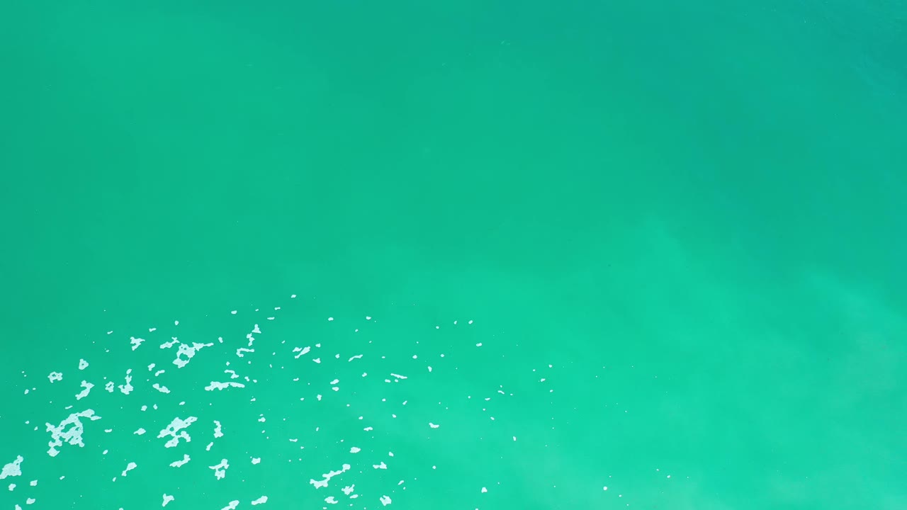 Overhead Los Cabos ocean surface with bright turquoise water, small white foam clusters across the lower portion, and no visible people.
