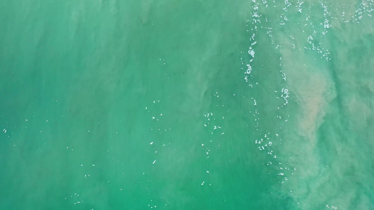 Straight-down aerial frame over green Los Cabos ocean water, where bright sparkles and cloudy sand shapes sit below the surface with no visible people.