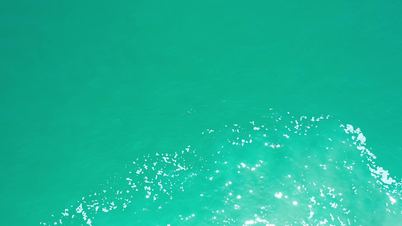 Overhead Los Cabos ocean frame with teal water filling the view and small white reflections gathered along the lower portion, with no visible people.