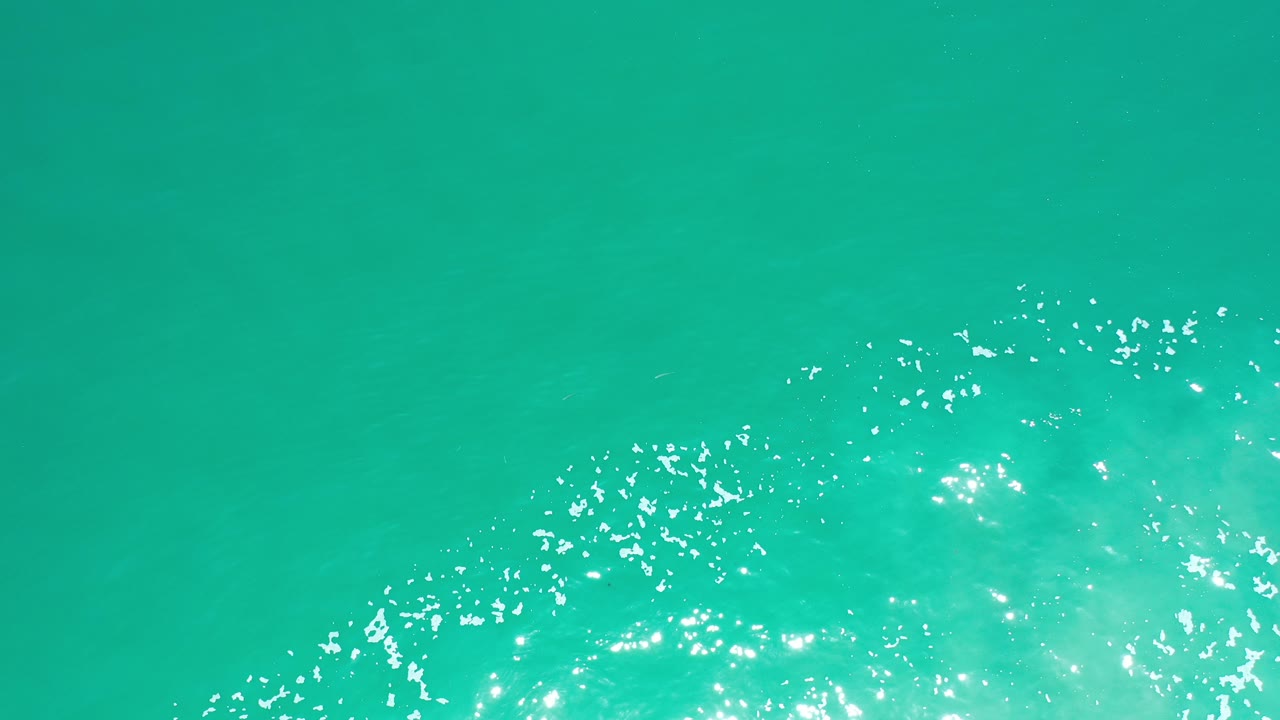 Overhead view of bright turquoise green sea in Los Cabos, Mexico, with sparkling sunlight clustered near the lower edge and no visible people.