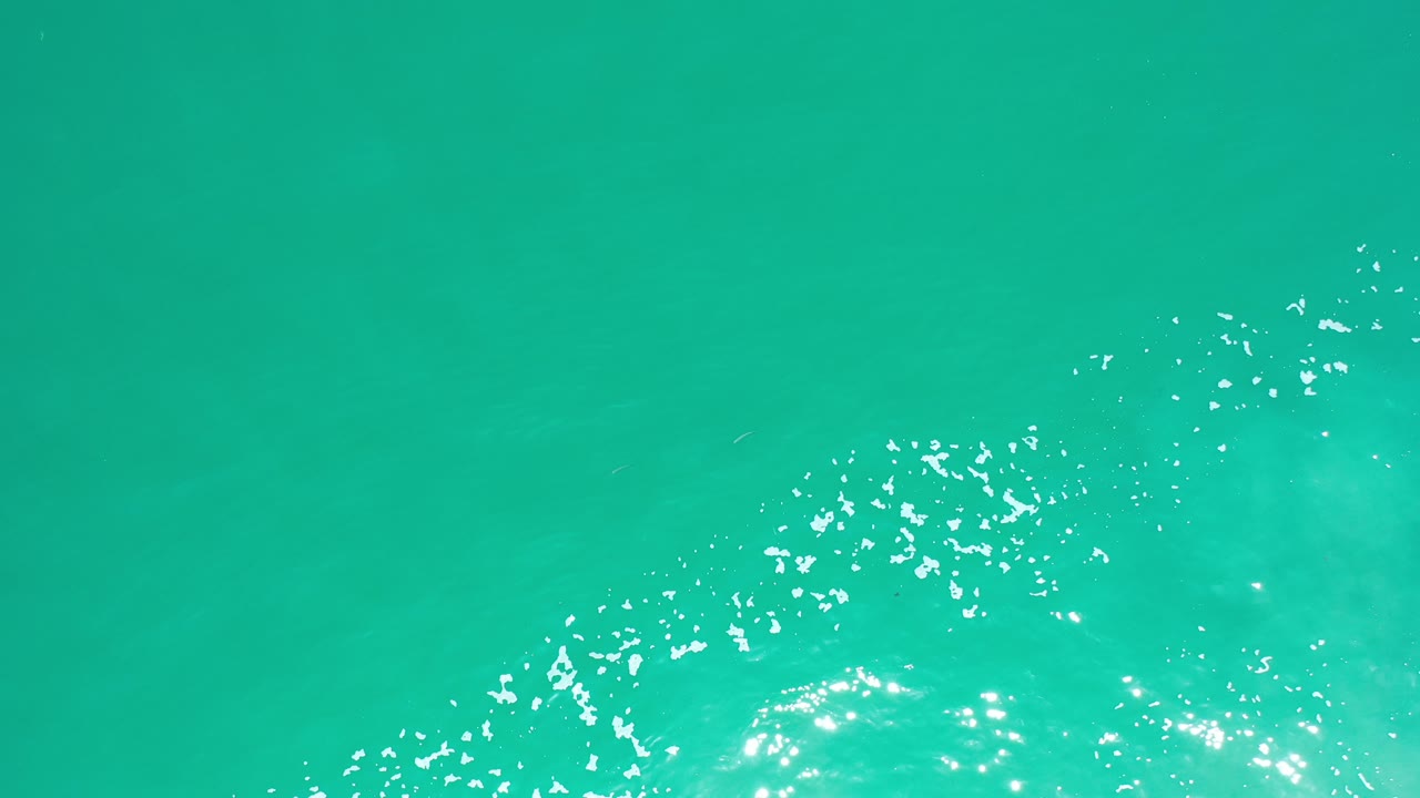 Top-down aerial view of greenish-blue water in Los Cabos, Mexico, with bright white foam clustered along the lower edge and no visible people.