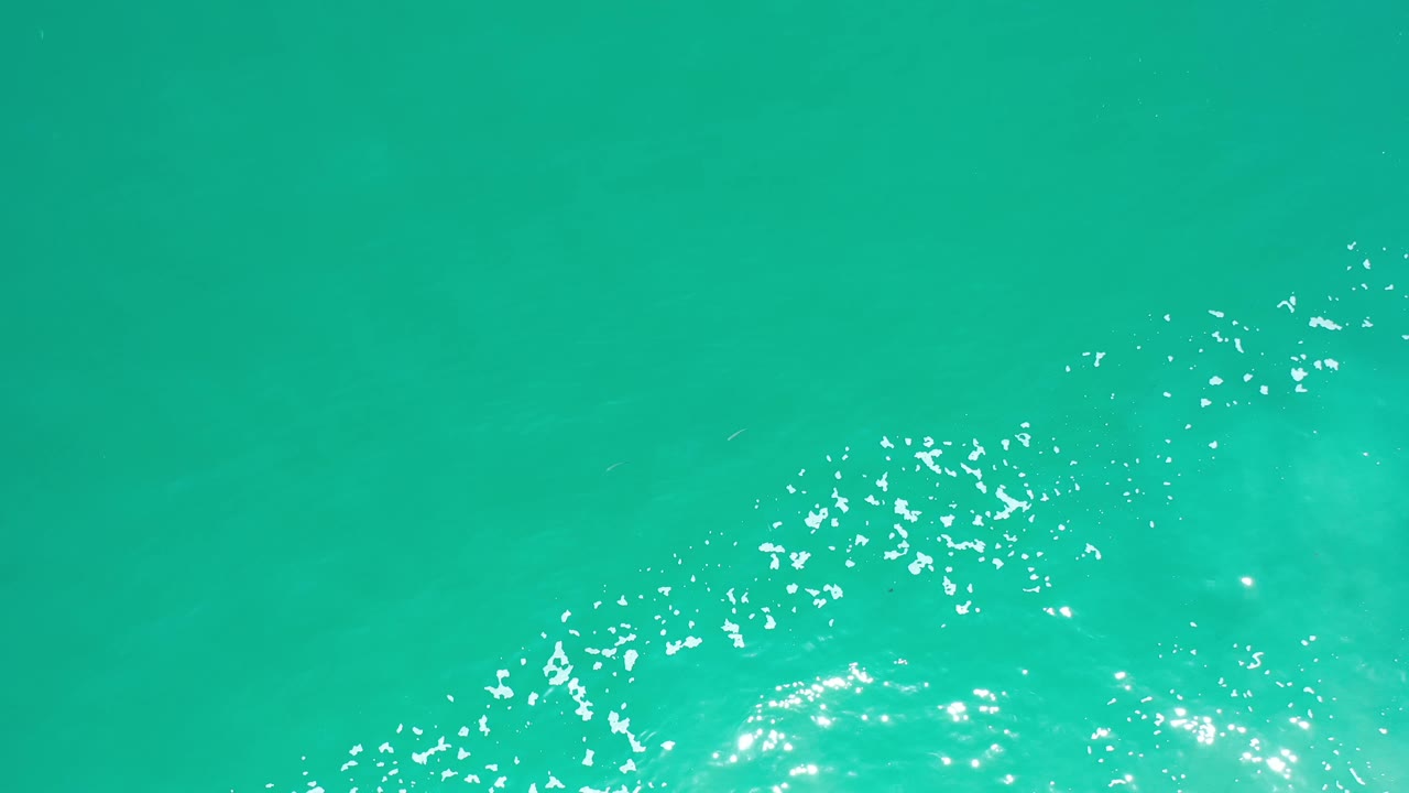 Overhead Los Cabos water frame with a diagonal band of white foam and glare crossing the lower portion, with no visible people or boats.