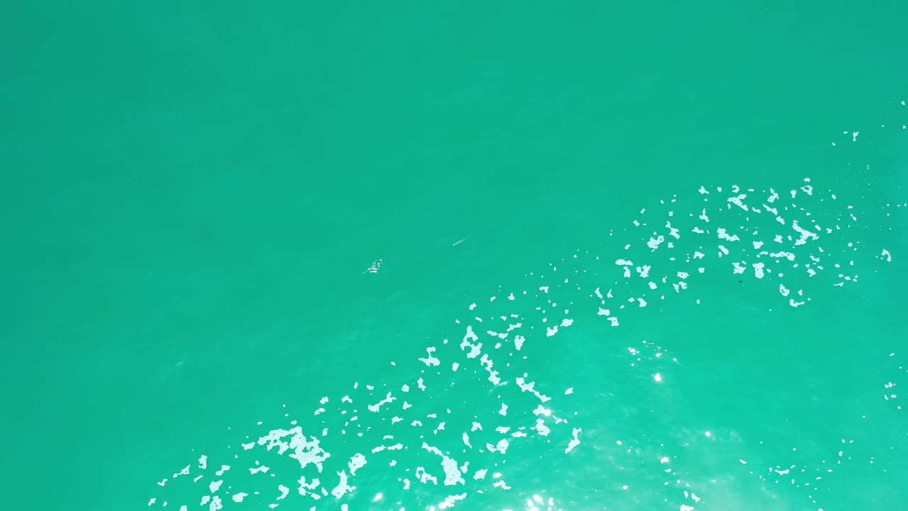 Aerial frame over Los Cabos water with a curved patch of white foam across the lower half and small bright glints on the greenish blue surface, no visible people.