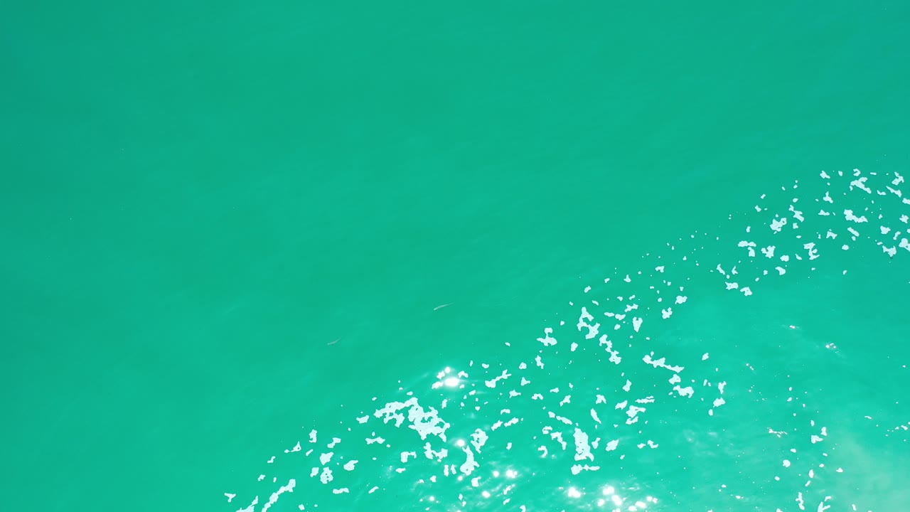 Overhead view of turquoise seawater in Los Cabos, Mexico, with a band of white foam entering from the lower left and no visible people.