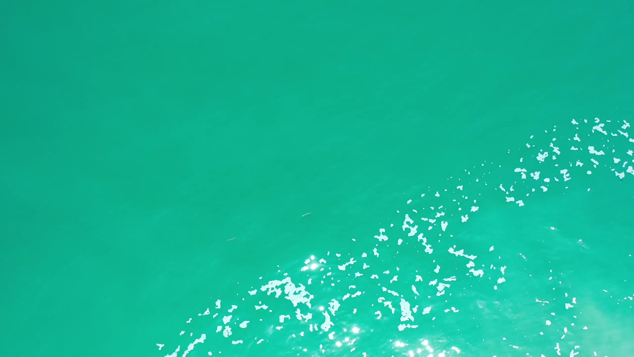 Aerial-looking view of aqua seawater in Los Cabos, Mexico, with scattered white foam and bright reflections concentrated along the lower edge, no visible people.