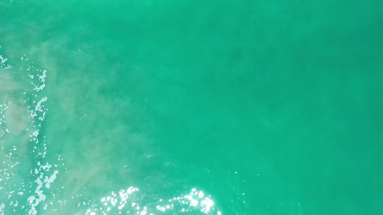 Overhead view of green-blue ocean water in Los Cabos, Mexico, with bright sun glints near the lower edge, faint pale swirls, and no visible people.