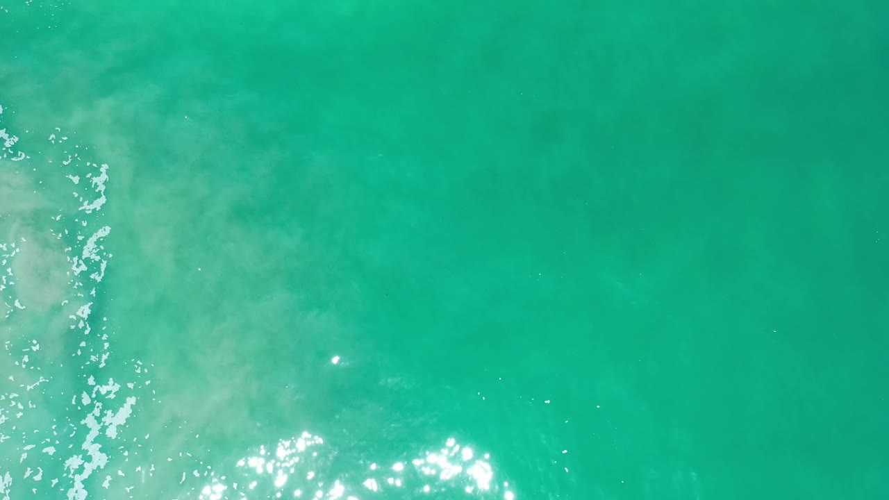 Aerial water surface in Los Cabos, Mexico, with a vertical band of foam and sun sparkle along the left side, cloudy pale water texture, and no visible people.