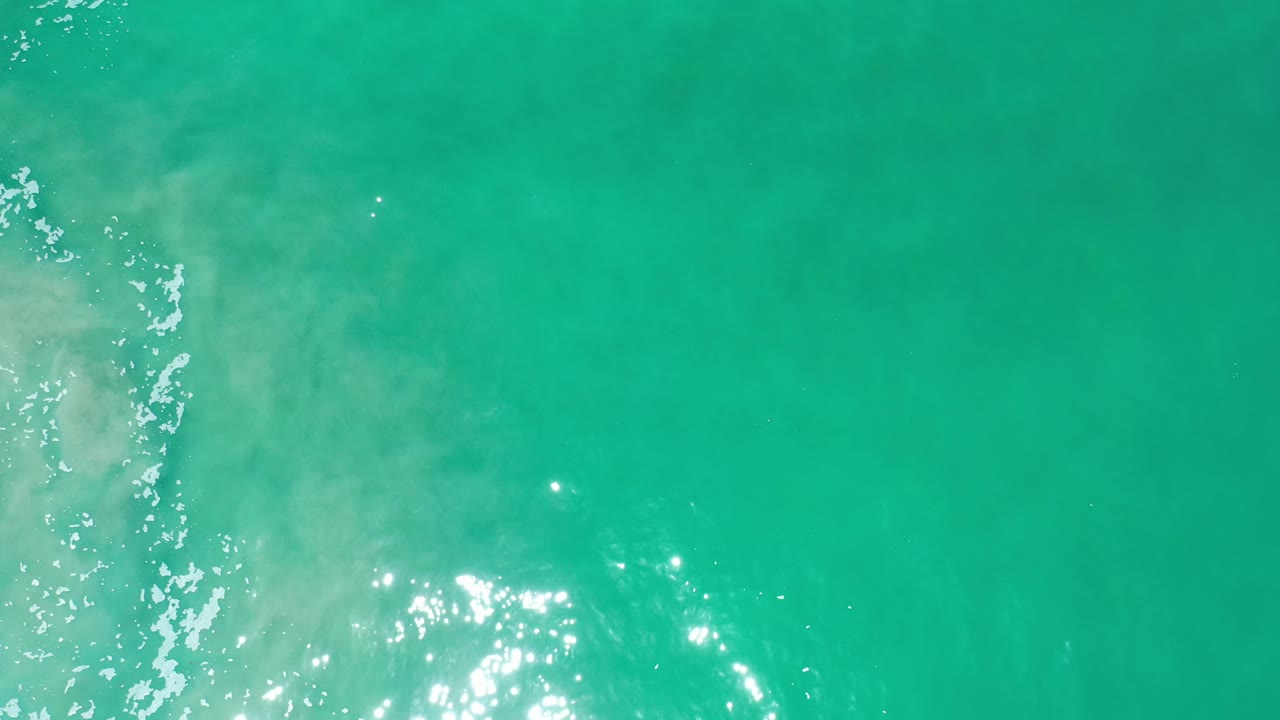 Overhead turquoise water with foam at left and bright glare near bottom.