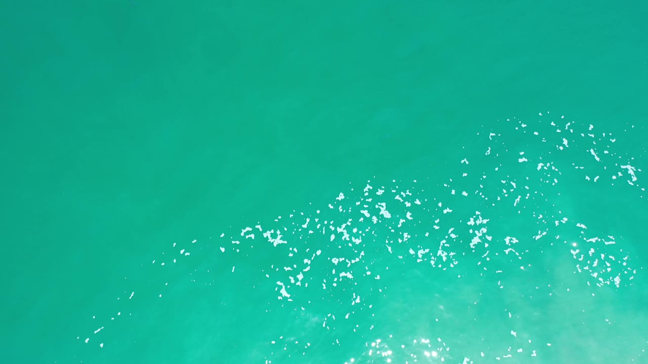 Overhead Los Cabos ocean frame with green-blue water and a broader arc of white surface sparkle near the lower center, with no visible people.