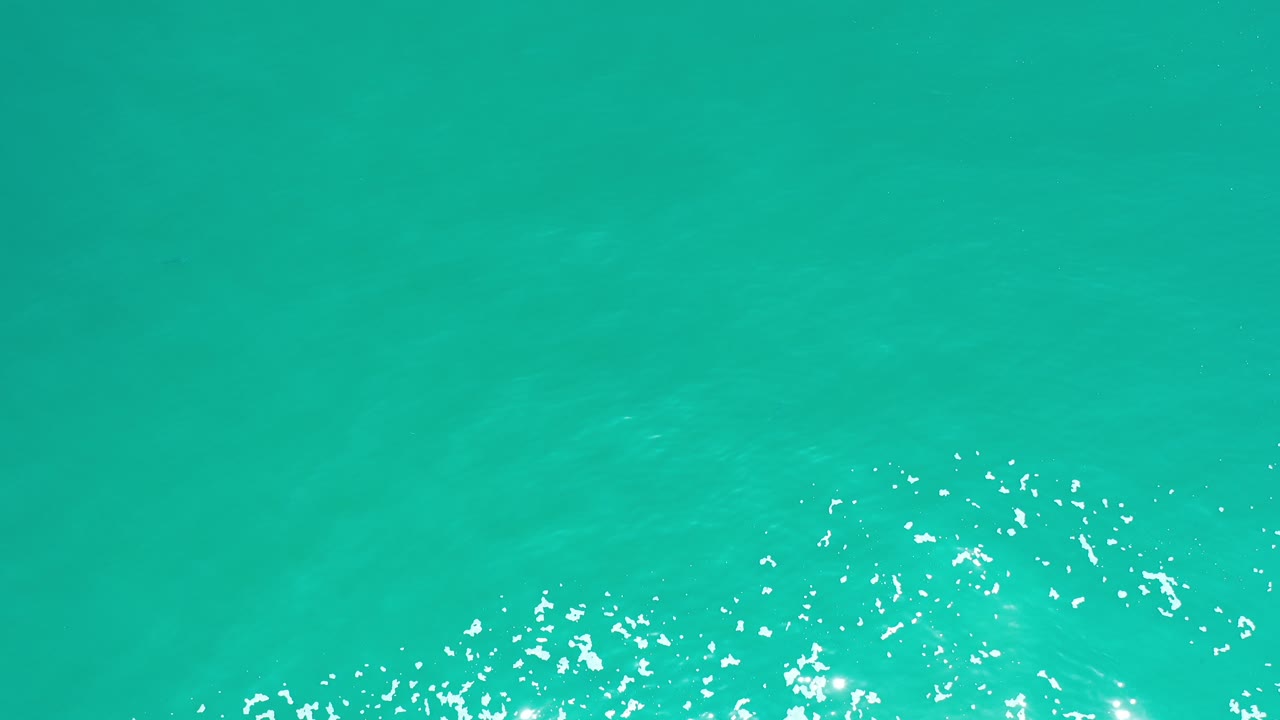 Overhead view of turquoise seawater in Los Cabos, Mexico, with scattered white foam and sun sparkle low in the frame, no visible people.