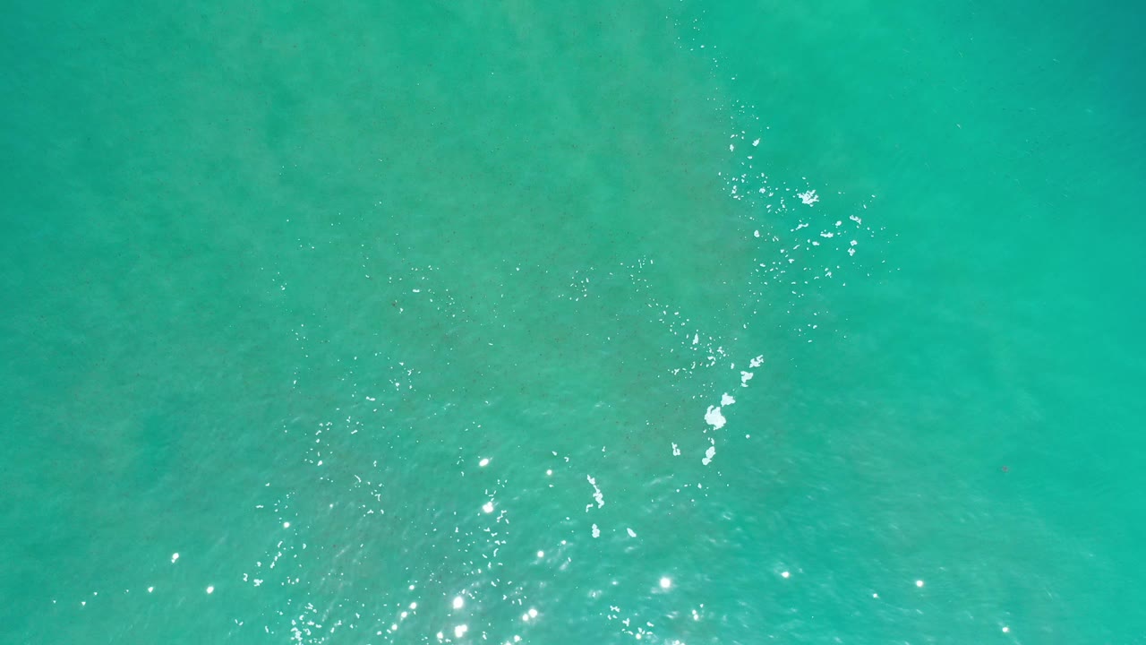 Overhead view of green-blue Los Cabos ocean water with small sun reflections, faint foam marks near the lower left, and no visible people.