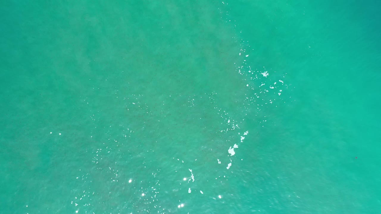 Green-blue ocean surface in Los Cabos, Mexico, with a narrow vertical foam streak and scattered bright reflections, no visible people.