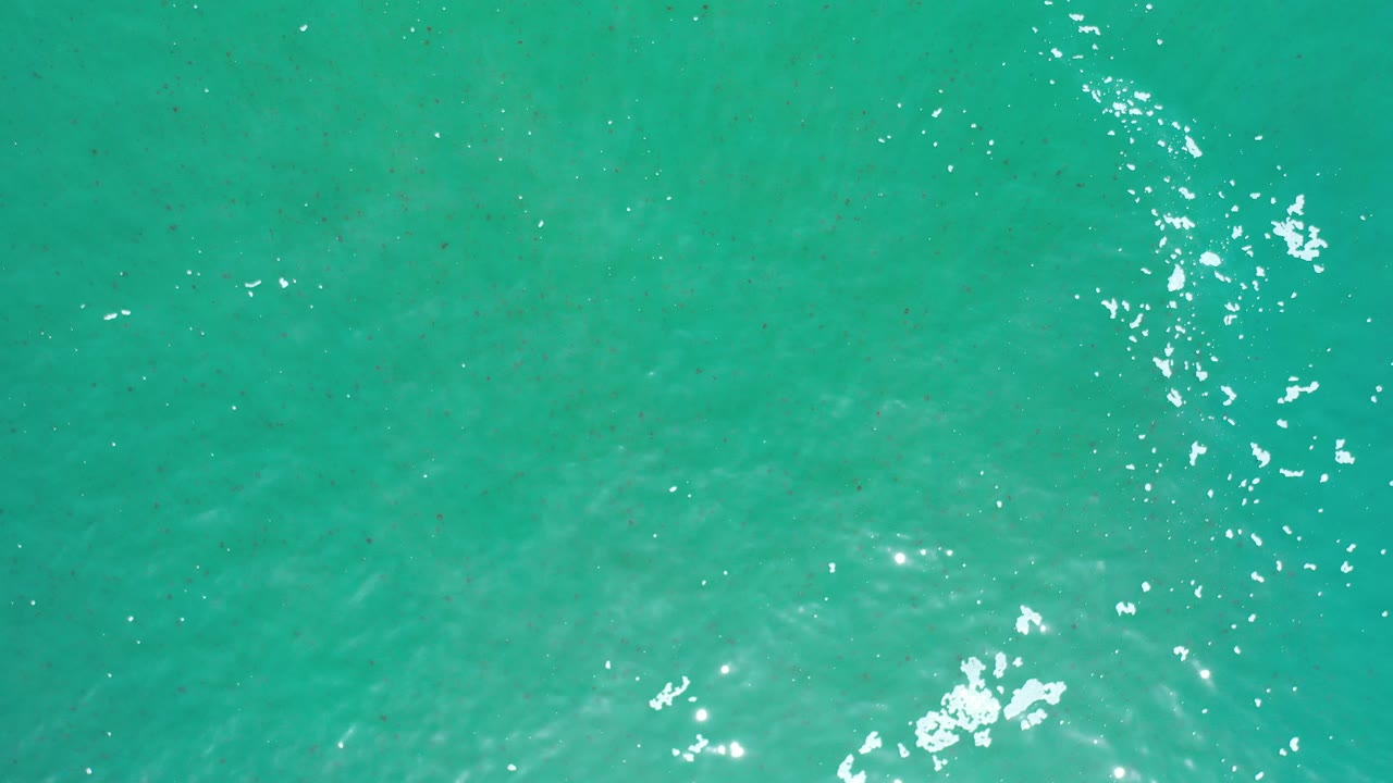 High view of turquoise ocean water in Los Cabos, Mexico, with scattered bright reflections near the upper right and no visible people.