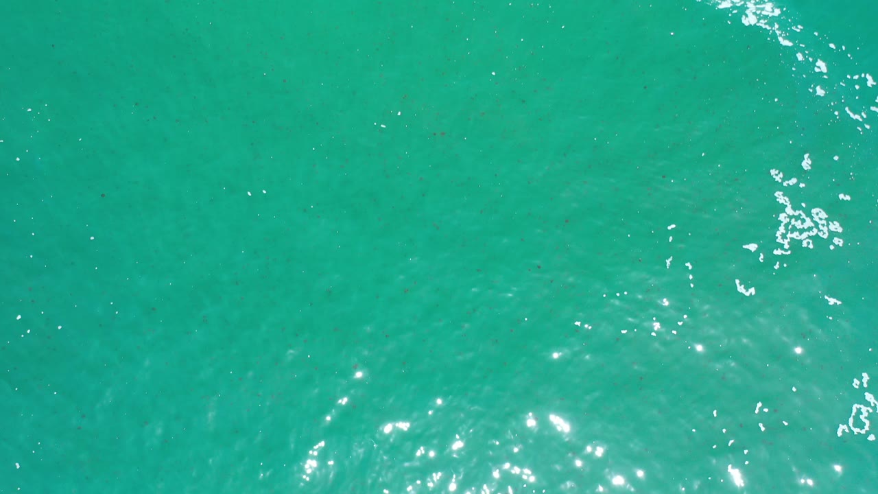 Overhead view of turquoise ocean water in Los Cabos, Mexico, with scattered white foam and bright sun reflections, no visible people.