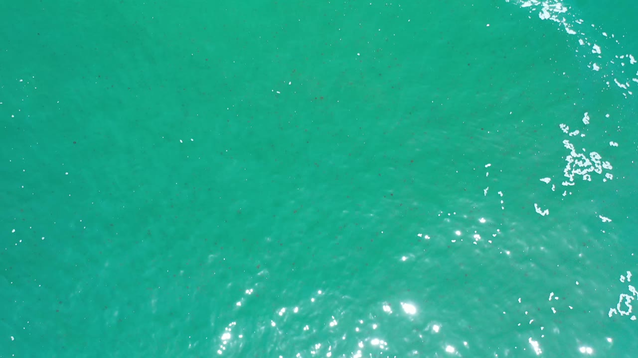 Overhead Los Cabos ocean surface with blue-green water, a diagonal cluster of white foam, and small points of reflected sunlight, no visible people.