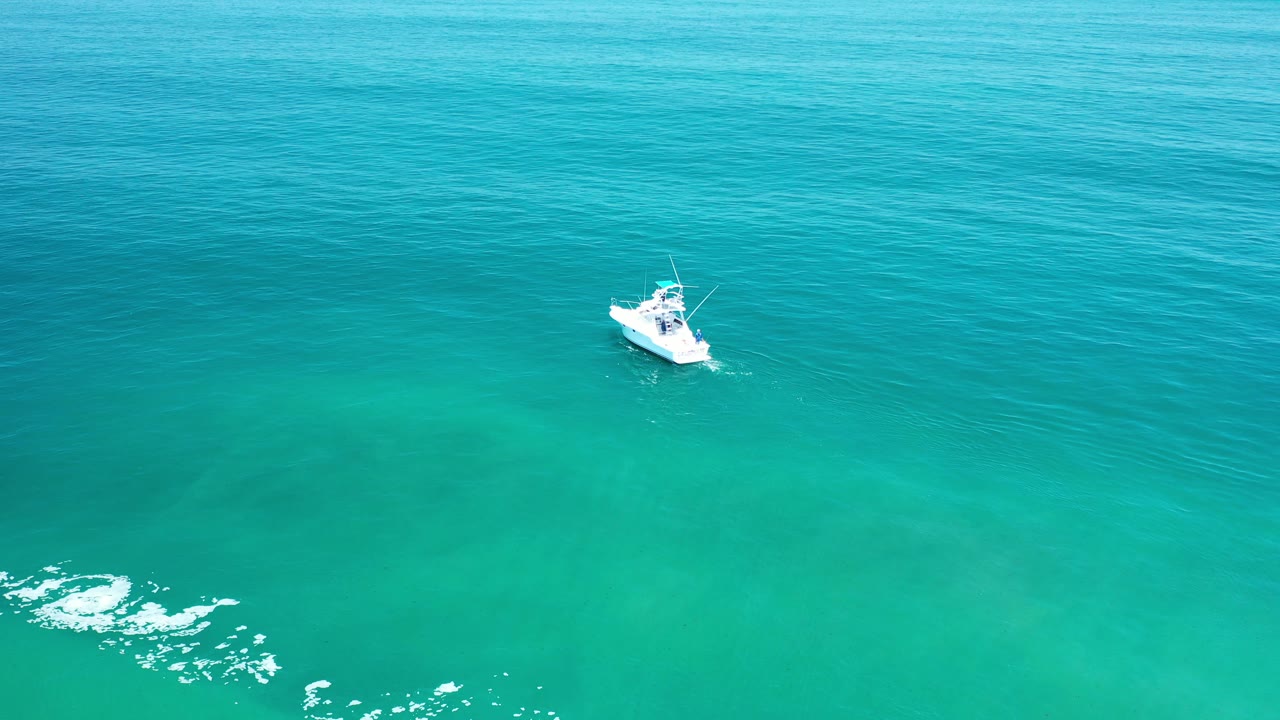 Aerial view of a small white boat on teal water in Los Cabos, Mexico, with bright surface glare low in frame and no visible people.