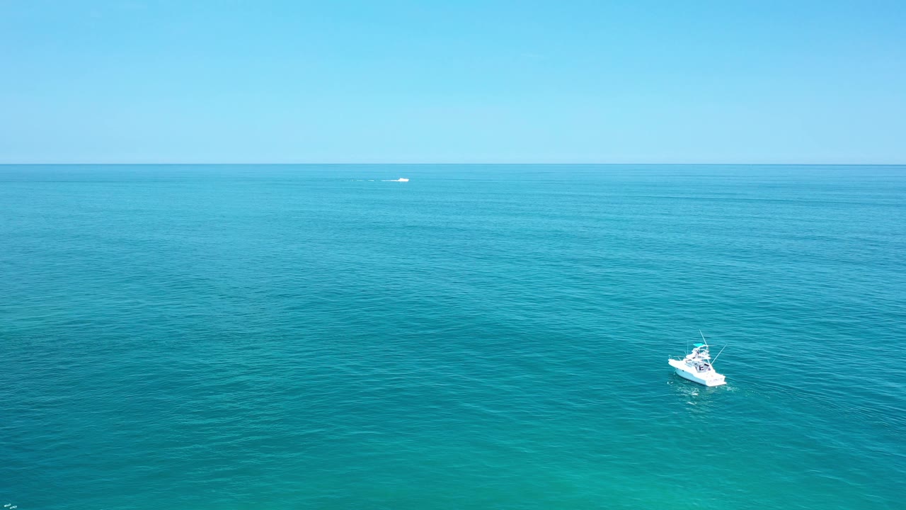 Aerial view in Los Cabos, Mexico of a small white boat on blue-green ocean below a clear sky, with no visible people.