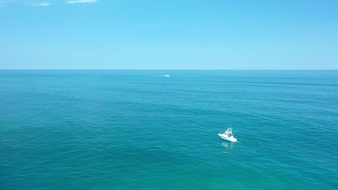 Aerial Los Cabos ocean frame with the small white boat slightly right of center, faint distant water traffic near the horizon, and no visible people.