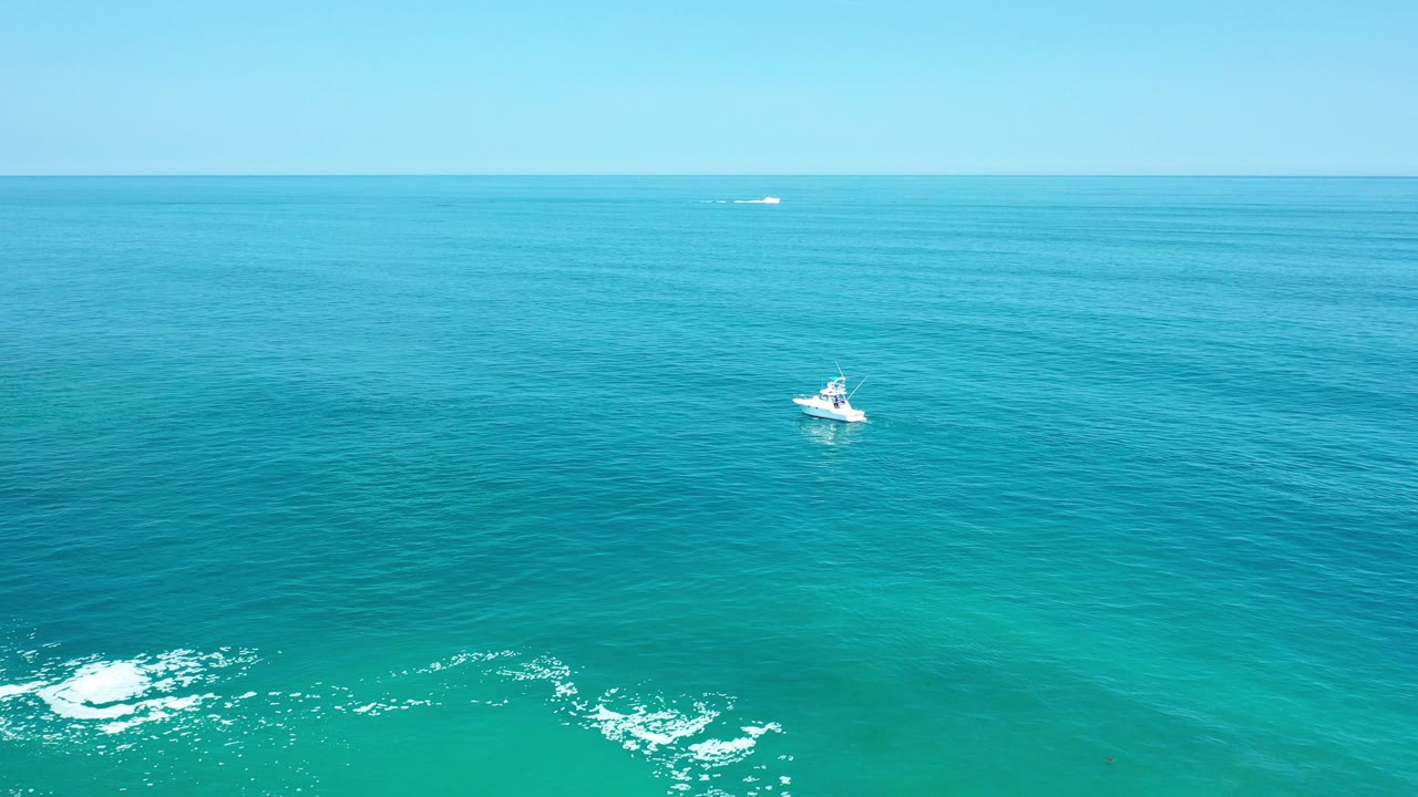 A small white boat floats off Los Cabos, Mexico, with turquoise water, a pale horizon, low surf at the bottom edge, and no visible people.