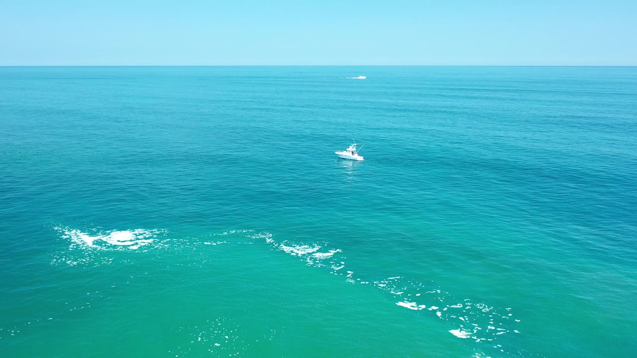 Aerial view in Los Cabos, Mexico, showing a small white boat beyond scattered white foam on turquoise water, with a level horizon and no visible people.