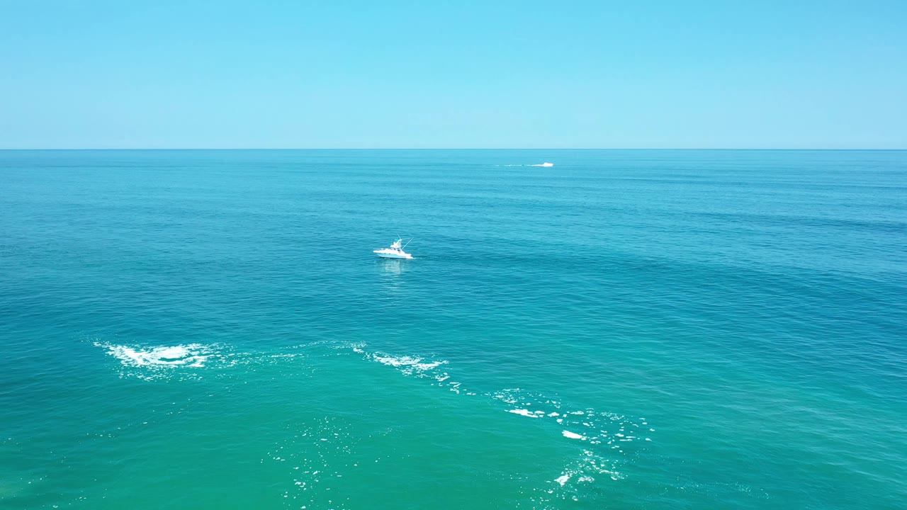 Aerial ocean frame from Los Cabos, Mexico, showing the small white boat near the center, a broad blue water field, faint distant vessels on the horizon, and no visible people.