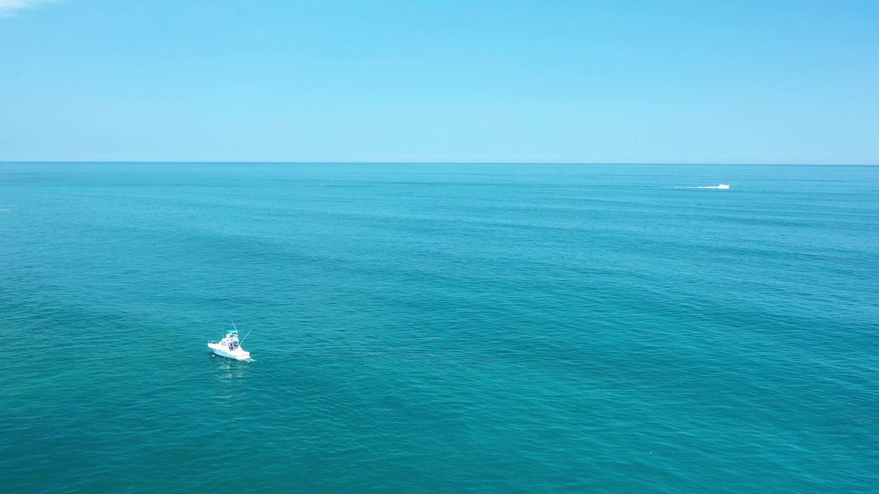 Wide aerial view in Los Cabos, Mexico, with a small white boat low left on turquoise green water, a level horizon, bright daylight, and no visible people.