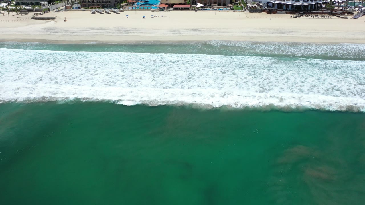 Overhead view of the Los Cabos shoreline with green water below, a wide band of breaking white surf, pale sand above, distant beachfront buildings, and no visible people.