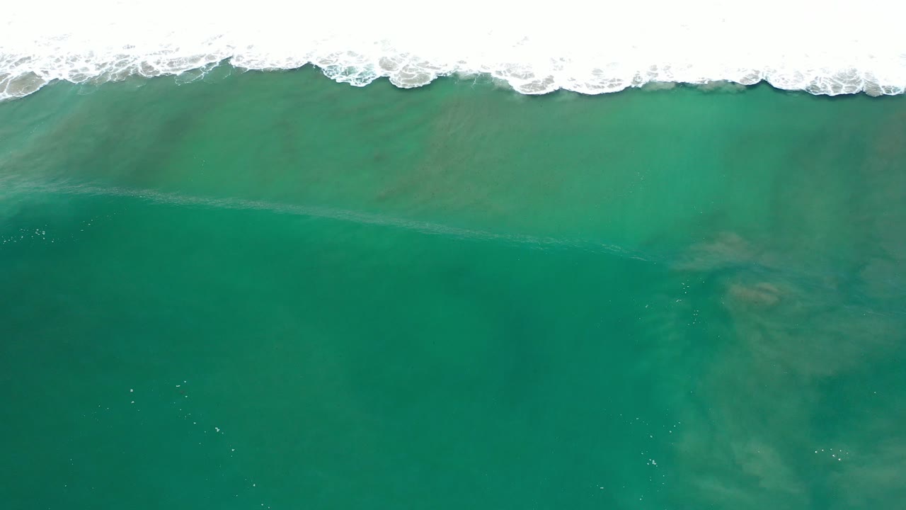 Overhead view of turquoise ocean water in Los Cabos with white surf along the top edge, faint underwater patches, and no visible people.