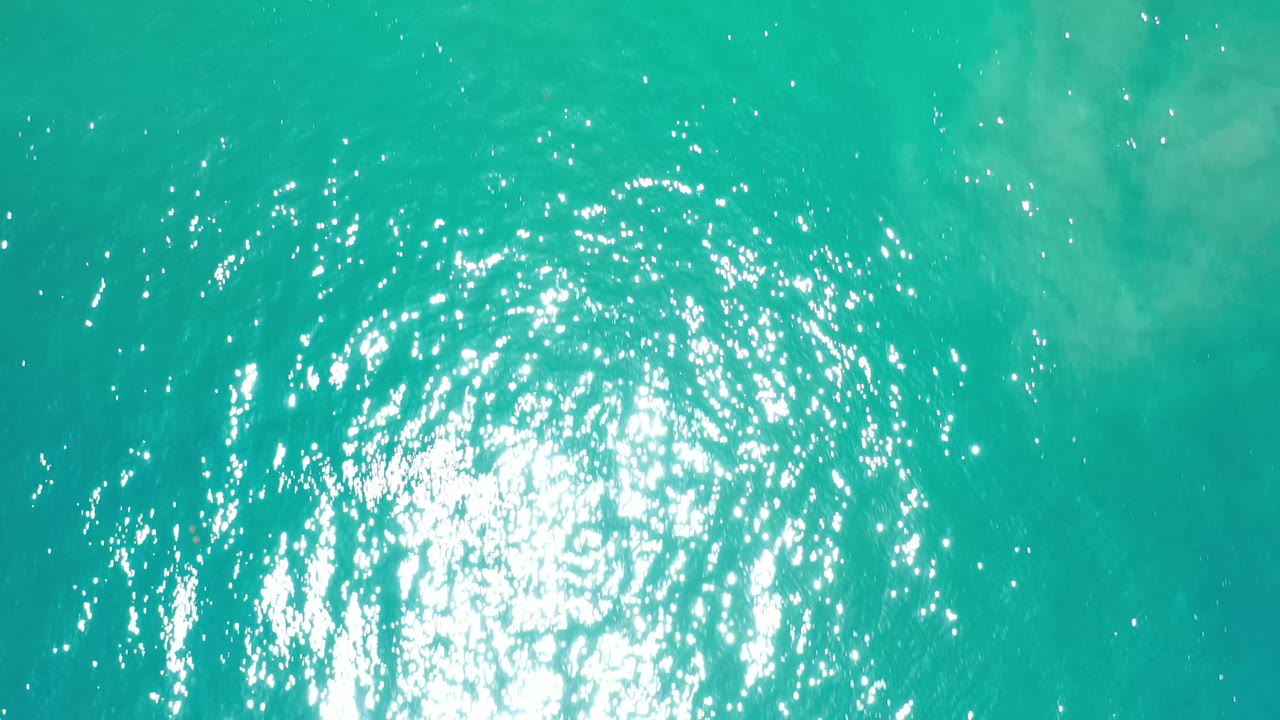 Overhead Los Cabos ocean frame with turquoise water, a centered patch of white glare, faint underwater color changes, and no visible people.