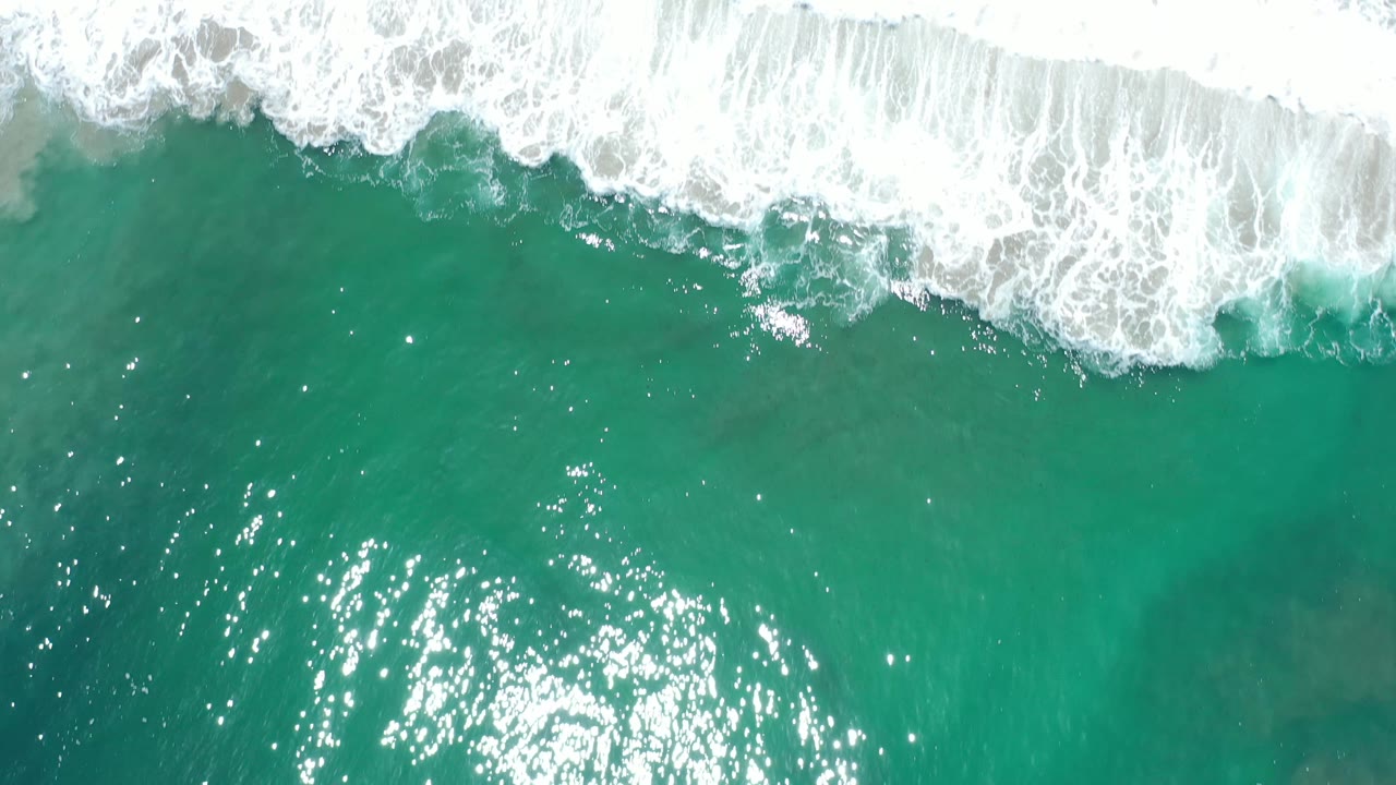Top-down Los Cabos ocean frame with bright sun reflections in teal water and white foam breaking across the top, with no visible people.