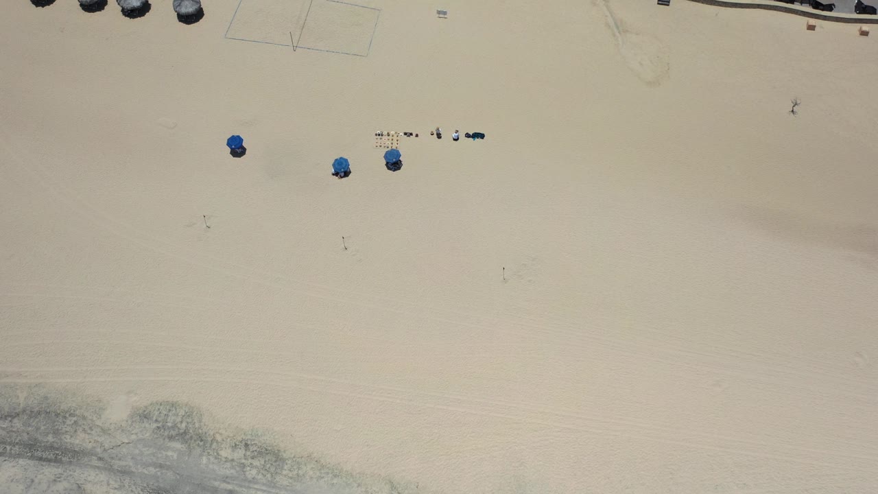 High overhead frame in Los Cabos, Mexico showing mostly pale beach sand, a few blue umbrellas, small beachgoers, and resort shade structures along the upper edge.