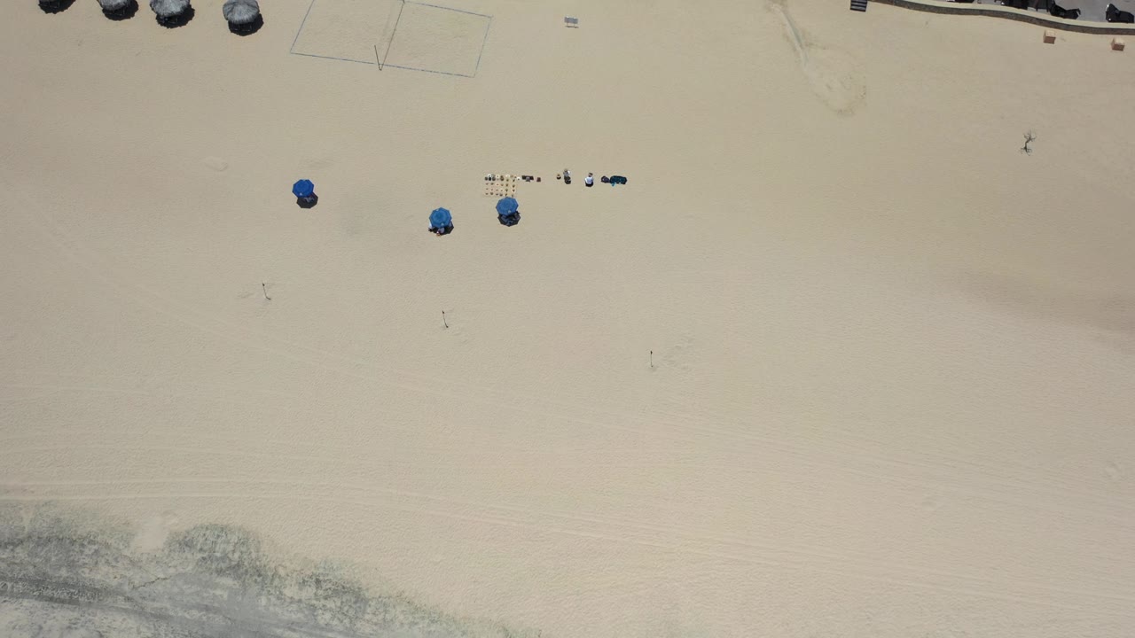 Overhead Los Cabos beach frame with broad open sand, scattered blue umbrellas, tiny visible visitors, faint beach court lines, and resort shade structures near the top.