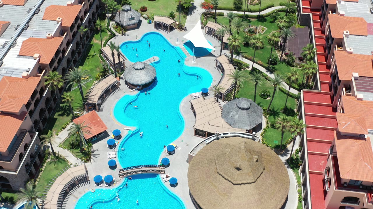 Aerial view of turquoise resort pools beside orange-roofed hotel buildings.