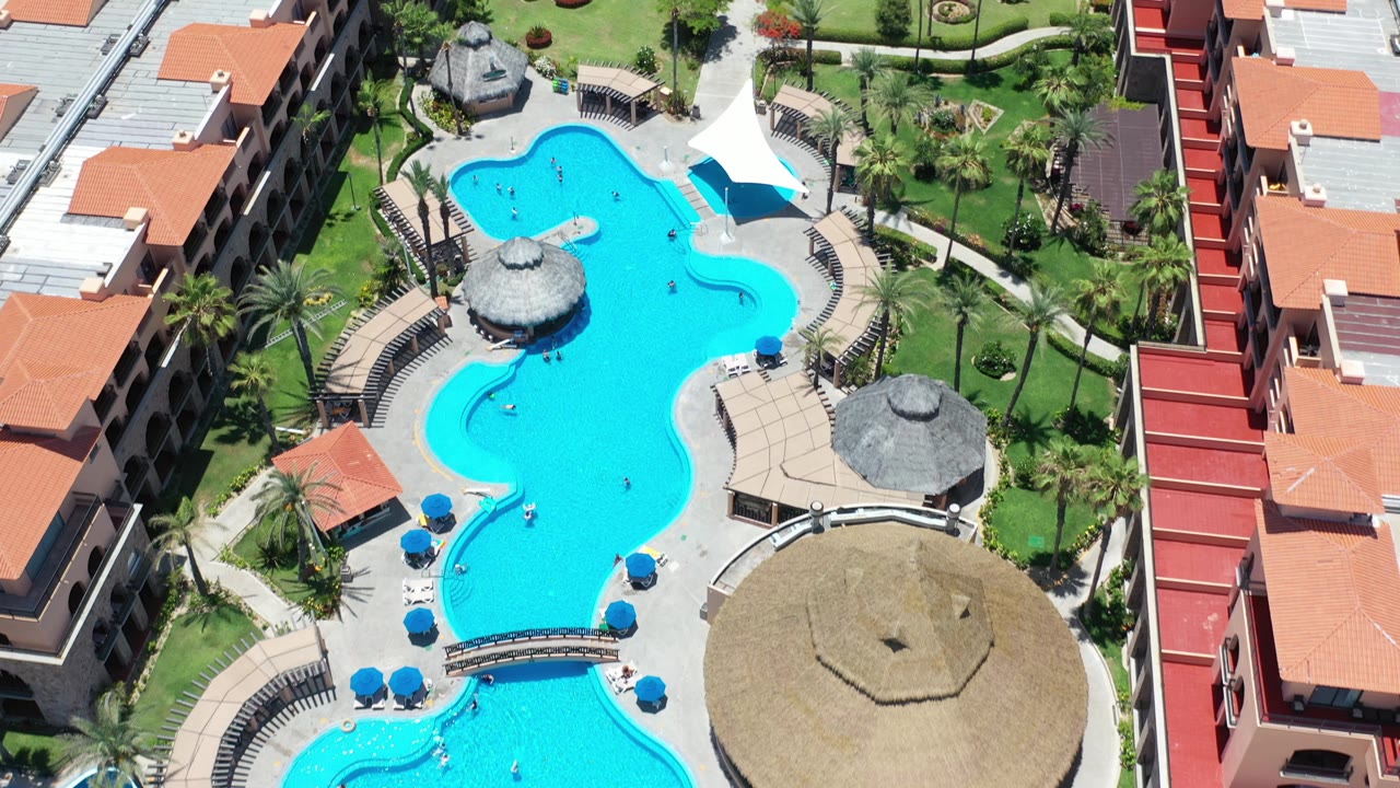 High aerial frame of the Los Cabos resort grounds, showing the pool complex, curved walkways, lawn areas, orange-roofed buildings, palapas, and no visible people.